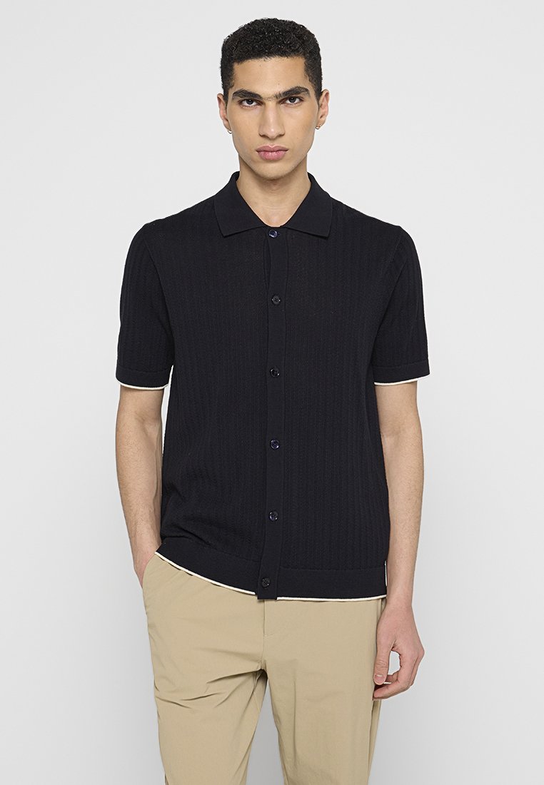 Granatowa koszula zapinana na guziki z krótkim rękawem o prążkowanej fakturze, z kołnierzykiem i kontrastowym jasnym akcentem u mankietu rękawa.