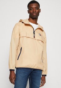 Lys beige, let jakke med halv-lynlås fortil, justerbar hætte og marineblå detaljer. Har en lynlås-lomme på siden og logo.