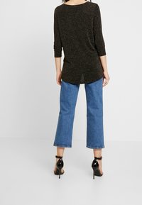 Schwarzes, glitzerndes Langarmtop mit abgerundetem Saum, kombiniert mit weit geschnittenen blauen Jeans und schwarzen High-Heels.