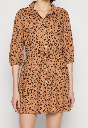 Kobieta ubrana w beżową sukienkę na guziki w czarne cętki lamparta, z rękawami za łokieć, kieszeniami na piersiach i paskiem wiązanym w talii.