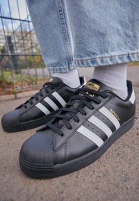 Svarta lädersneakers med tre vita ränder, rund tån och texturerad gummisula. Guld "adidas"-logotyp på tungan och sidan.