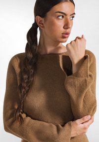 Brauner gerippter Strickpullover mit weiten Ärmeln, entspannter Halslinie und weicher Textur. Haare zu einem Zopf gestylt, Hände an der Taille verschränkt.