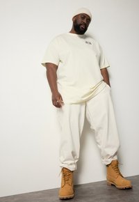 Světlě žluté oversized tričko, krémové volné kalhoty a hnědé šněrovací boty. Tkanina vypadá měkce; na tričku je jemný detail loga.