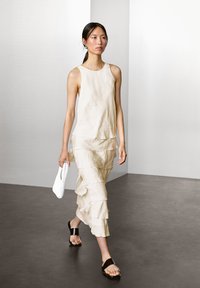 Beiges Leinenkleid mit schichtweisen Rüschen und rundem Ausschnitt. Kombiniert mit schwarzen Sandalen und einer weißen geflochtenen Clutch. Einfaches, strukturiertes Design.