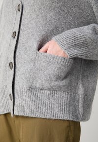 Main dans la poche avant d'un cardigan gris en tricot avec des boutons marron, porté sur un pantalon marron.