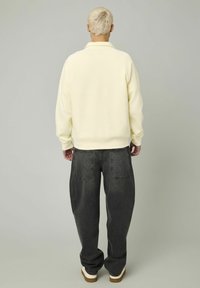 Personne avec des cheveux blonds courts, portant un sweatshirt crème, un jean noir ample et des chaussures blanches, se tenant devant un fond gris uni.