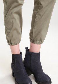 Zwarte enkellaarzen met een hoge blokhak, een gladde leren textuur, zijritssluiting, gecombineerd met olijfgroene tapered broeken met elastische manchet.