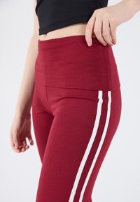 Leggings de sport rouges avec une taille haute et deux bandes blanches sur les côtés, fabriqués à partir d'un tissu lisse et extensible.