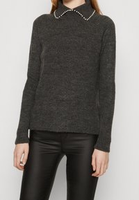 Maglione grigio scuro lavorato a maglia con finitura testurizzata e colletto piatto ornato da dettagli simili a perle, indossato sopra pantaloni attillati neri.