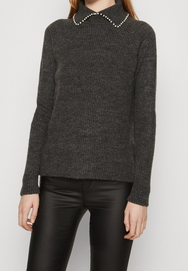 Maglione grigio scuro lavorato a maglia con finitura testurizzata e colletto piatto ornato da dettagli simili a perle, indossato sopra pantaloni attillati neri.