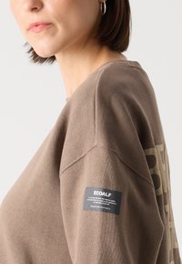 Brun genser med ribbestrikket rund hals, med trykt logo og et stoffmerke på ermet. Materialet virker mykt.