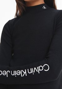 Maglietta nera a coste con maniche lunghe e collo alto. Presenta il testo bianco "Calvin Klein" sulla manica sinistra e un logo argentato sul collo.