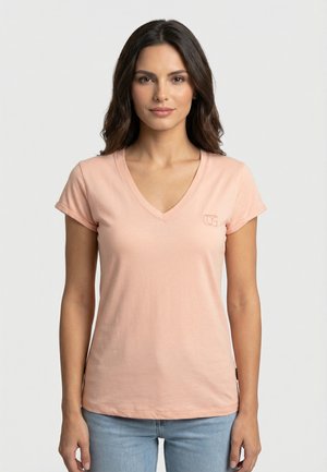 Femme aux longs cheveux foncés portant un T-shirt rose clair à col en V et un jean bleu, debout devant un fond blanc uni.