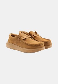 Des chaussures slip-on en suede marron avec un bout rond, des détails cousus et une semelle en caoutchouc, présentant un accent de lacet et un logo sur le côté.