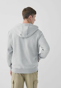 Sudadera gris con capucha ajustable, puños y dobladillo acanalados. Textura suave, diseño casual y corte relajado. El fondo es completamente blanco.