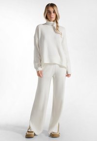 Witte gebreide coltrui gecombineerd met bijpassende wijde broek, beide met een zachte textuur en een ontspannen pasvorm. Verstevigd met zij-slits.