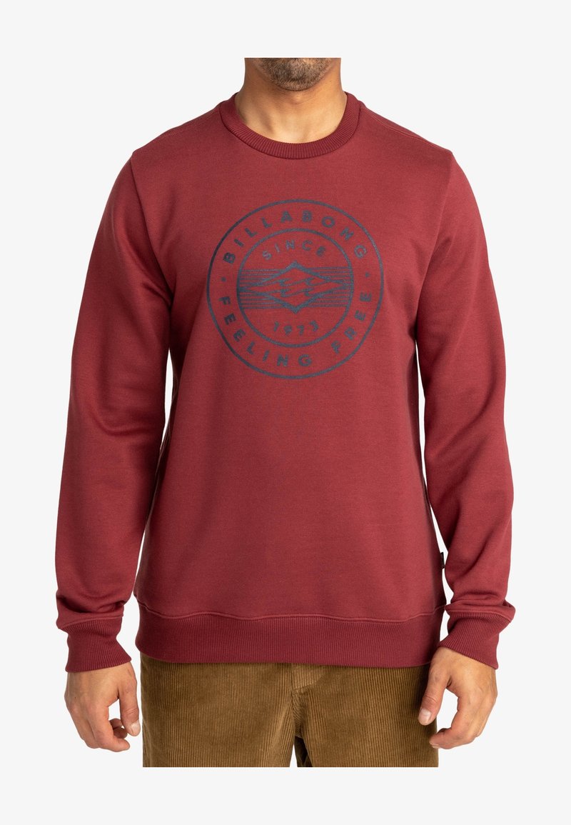 Homme portant un sweat-shirt à manches longues bordeaux avec un logo rond Billabong et le texte "Since 1973 Feeling Free" sur la poitrine.
