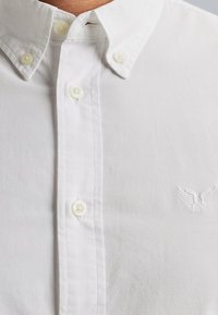 Camisa blanca de botones con cuello puntiagudo, que presenta cuatro botones blancos y un sutil logo bordado de ala en el lado izquierdo del pecho.