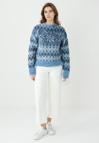 Gestrickter Pullover in verschiedenen Blautönen mit geometrischem Muster, mit Rundhalsausschnitt und gerippten Bündchen, kombiniert mit weißen Hosen mit weitem Bein.