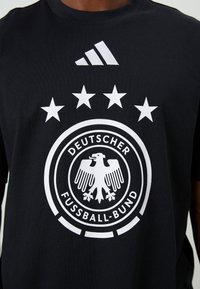 T-shirt preta de mangas curtas com um grande emblema circular branco com uma águia e cinco estrelas, e um logo branco da Adidas no peito.