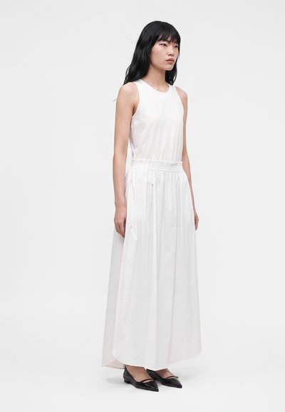 Femme aux longs cheveux noirs portant une robe maxi blanche sans manches et des ballerines noires pointues, debout devant un fond clair uni.