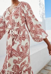 Kobieta w białej sukience z rdzawoczerwonym nadrukiem paisley, z bufiastymi długimi rękawami i związanym paskiem w talii, stojąca obok białej ściany pod niebieskim niebem.