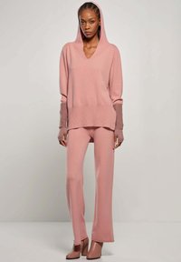 Różowy dzianinowy sweter z kapturem, dekoltem w serek i prążkowanymi mankietami, w połączeniu z dopasowanymi spodniami. Gładka tekstura i luźny krój. Buty na obcasie.