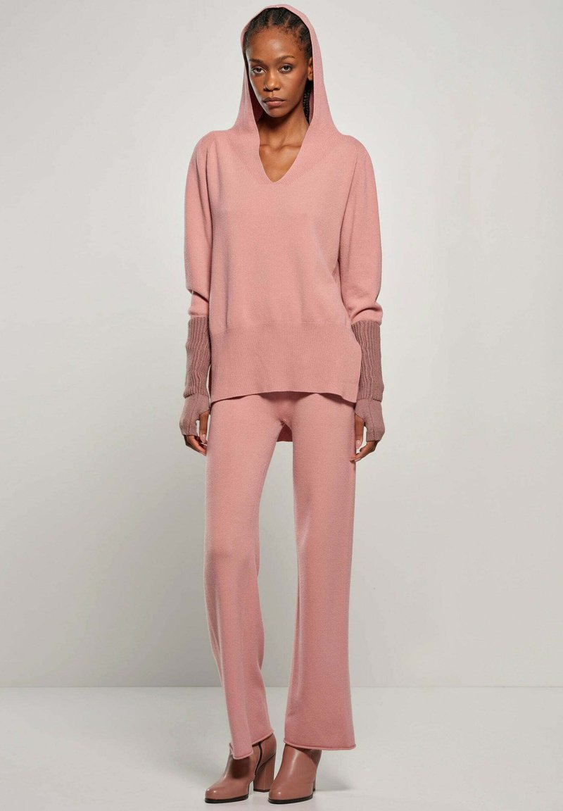 Różowy dzianinowy sweter z kapturem, dekoltem w serek i prążkowanymi mankietami, w połączeniu z dopasowanymi spodniami. Gładka tekstura i luźny krój. Buty na obcasie.