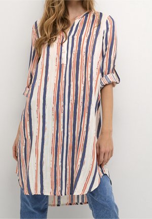 Vrouw draagt een losse, gestreepte tuniek met verticale lijnen in blauw, oranje en wit, gecombineerd met een blauwe spijkerbroek en opgerolde mouwen.