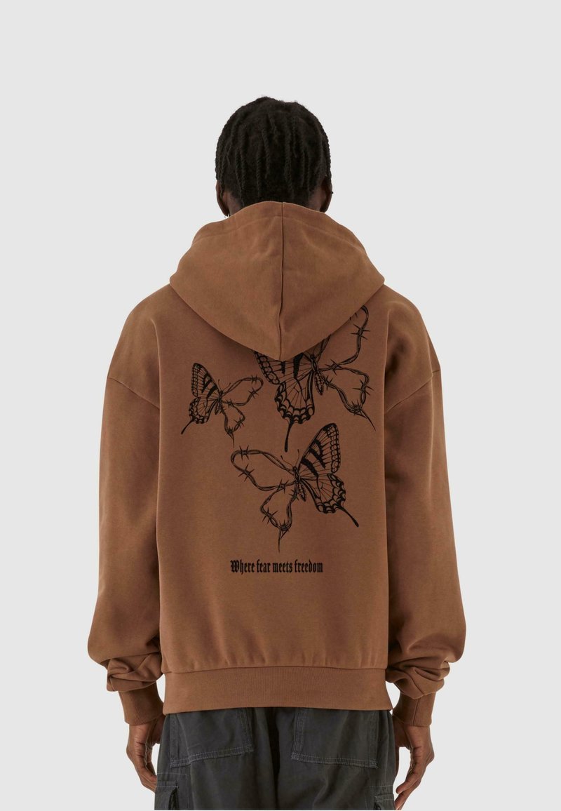 Personne aux cheveux foncés et bouclés portant un sweat à capuche marron avec un dessin de papillon noir et de fil barbelé, avec le texte « Où la peur rencontre la liberté » dans le dos.