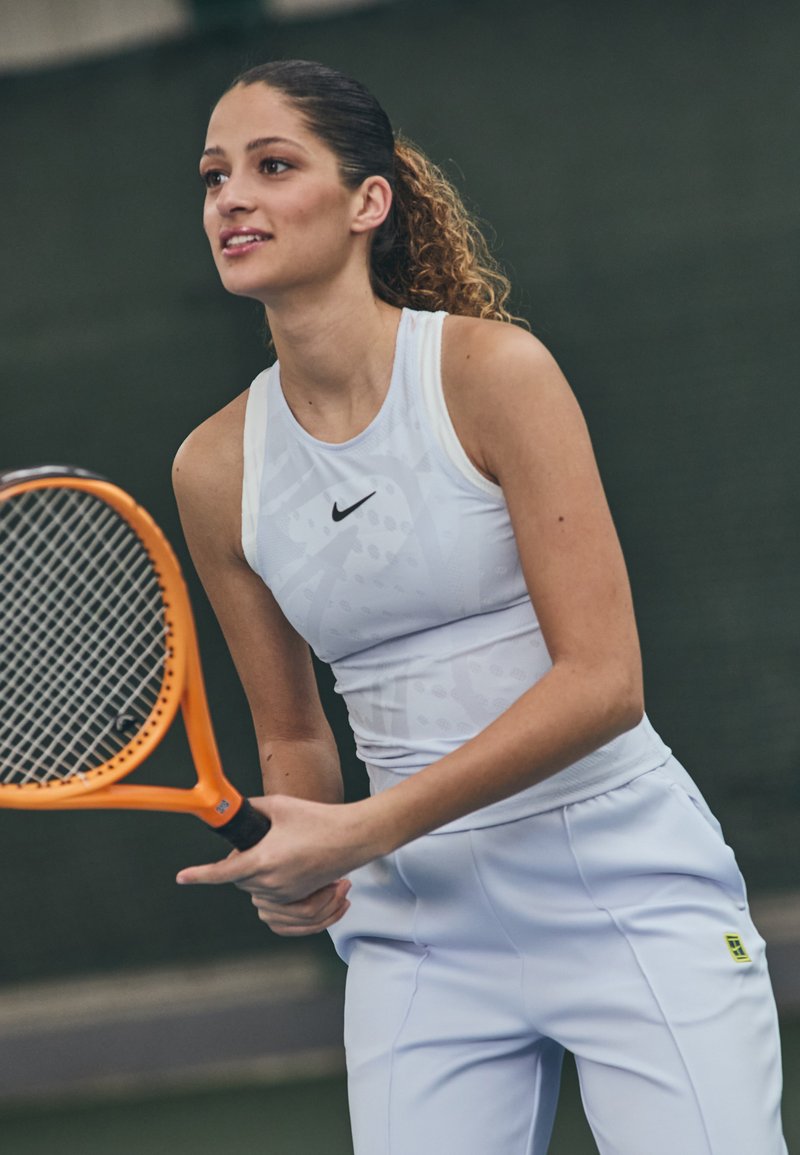Fehér sportfelső diszkrét mintázattal és fekete Nike logóval, világosszürke nadrággal párosítva. Narancssárga teniszütő a kezében.