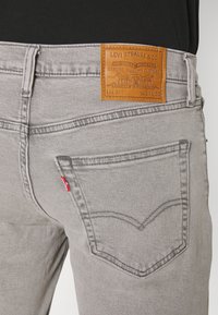 Vaqueros de mezclilla gris con un parche de cuero de Levi Strauss en la cintura, bolsillos traseros visibles y un acento de tab rojo en la parte izquierda.