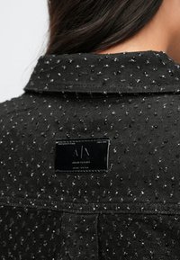 Čierna džínsová bunda s textúrovanou látkou s malými vypuklými bodkami, špicatým golierom a značkovou štítkom na zadnej strane.