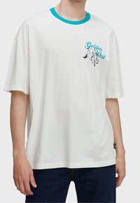 Camiseta blanca con cuello turquesa, pequeño gráfico de un vaquero montando un caballo desbocado, y texto "Golden West" en el lado izquierdo del pecho.