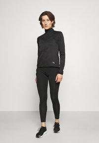 Schwarze Sportjacke mit hohem Kragen, Reißverschluss vorne und strukturierten Einsätzen. Kombiniert mit schwarzen Leggings und Schuhen, um einen eleganten Look zu kreieren.
