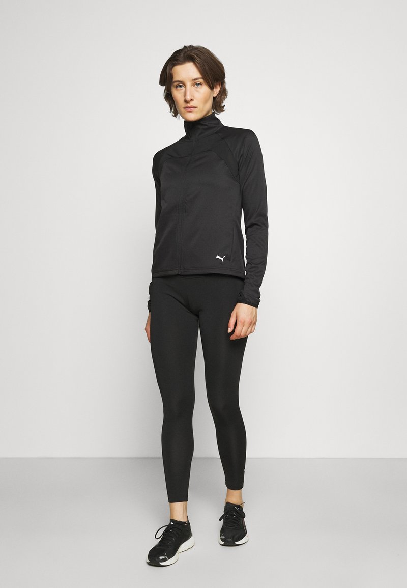 Schwarze Sportjacke mit hohem Kragen, Reißverschluss vorne und strukturierten Einsätzen. Kombiniert mit schwarzen Leggings und Schuhen, um einen eleganten Look zu kreieren.