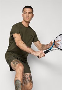 Olivengrønn trenings T-skjorte med korte ermer og logo, sammen med matchende shorts, med synlige tatoveringer på beina. Holder en tenniskølle.