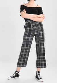 Černý top bez ramínek s řasením, spárovaný s kostkovanými kalhotami s širokým lemem v černé, šedé a bílé barvě, s knoflíkovým zapínáním v přední části. Černé tenisky.
