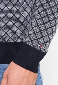 Pull en tricot avec un motif en losanges bleu marine et gris, des poignets côtelés et une petite étiquette logo avec des accents rouge, blanc et bleu sur la manche.