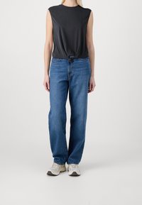 Top grigio senza maniche con un nodo all'orlo, abbinato a jeans dritti azzurri chiari, indossato con sneakers beige e grigie.