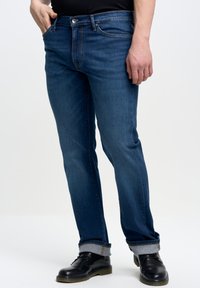 Vaqueros de mezclilla azul con corte recto, con cierre de botón, cinco bolsillos y puños doblados, combinados con zapatos de cuero negros.