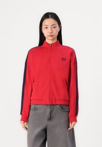 Giacca rossa con zip e accenti blu navy, colletto alto e due tasche laterali. Realizzata in tessuto morbido e con un piccolo logo sulla parte sinistra del petto.