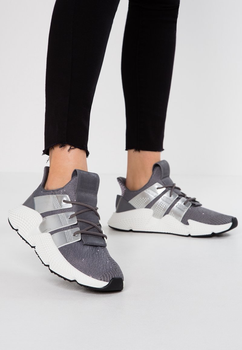 Grå sportskor med silverdetaljer och reflexband. Texturerad stickad ovandel och tjock vit mellansula, med en gummiyttersula.