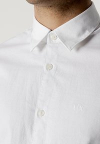 Camicia bianca in cotone con colletto a punta, dotata di chiusure a bottone discrete e un leggero logo ricamato nell'area della tasca sul petto.