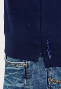 Navy blue cotton T-shirt with a straight hem and side split, featuring embroidered "U.S. Polo Assn" text on the bottom left. Worn over light blue denim jeans.