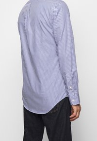 Mies, jolla on päällään pitkähihainen sinivalkoraitainen paita, joka on ulkona tummansinisistä farkuista, kuvattuna takaa.