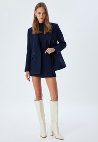 Marineblauw tweed blazer over een bijpassende rok, gecombineerd met glanzende witte kniehoge laarzen. Heeft een gestructureerd ontwerp en een dubbele knoopsluiting.