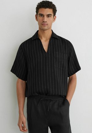 Man draagt een zwart shirt met korte mouwen met witte verticale strepen en zwarte broek met trekkoord, staande tegen een witte muur.
