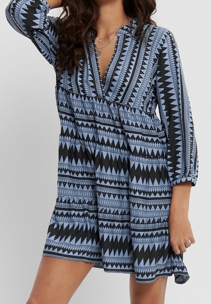 Femme portant une robe à motifs géométriques bleus et noirs, manches longues et col en V, accessoirisée avec des colliers argentés superposés.
