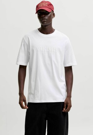 Dark-skinned person wearing a red cap, white t-shirt with "ORIGINALS" text, and loose black pants, standing against a plain light background.